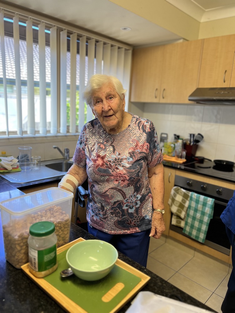 An older woman prepares food in her kitchen at home, with a mixing bowl and canisters on the bench.